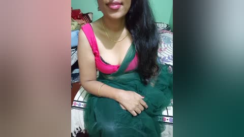 Media: Video of a South Asian woman with medium brown skin and long black hair, wearing a green sari with a pink blouse, sitting on a bed with patterned sheets.