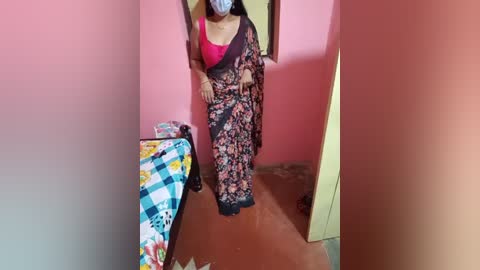 Media: A video of a woman in a floral saree with a pink blouse, standing in a small, pink-walled room with a colorful ironing board and iron.