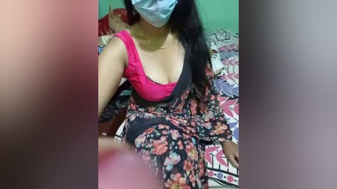 Media: A video of a South Asian woman in a pink bra, floral dress, face mask, and blue face shield, sitting on a bed with a colorful quilt, in a dimly lit room.