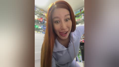 Media: A video of a young Asian woman with long, straight red hair and fair skin, wearing a light blue lab coat, smiling in a brightly lit pharmacy aisle filled with colorful products.