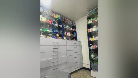 Video of a small, well-lit room with white cabinets filled with colorful medication boxes and a gray counter. The room is clean and organized, with a speckled beige tile floor.