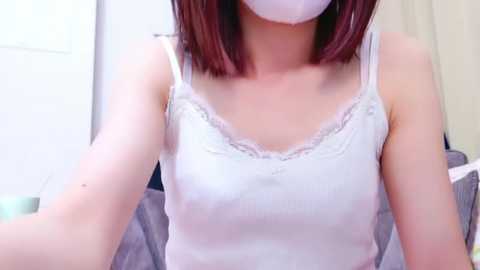 Media: A video of an Asian woman with shoulder-length brown hair, wearing a white lace-trimmed tank top, white face mask, and a white headband. She is indoors with a blurred background.