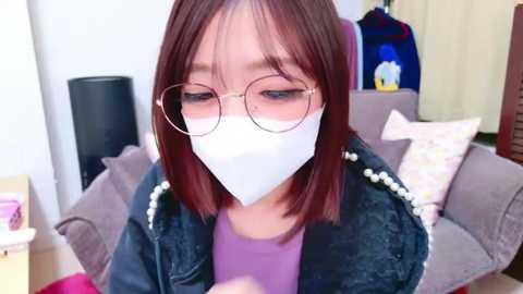 Media: Video of an East Asian woman with shoulder-length brown hair and glasses, wearing a white face mask, black jacket, and purple top, seated in a living room with gray couch, floral pillows, and a blue backpack.