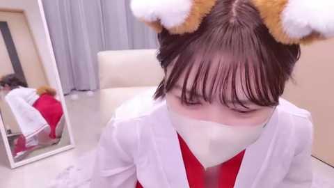 Media: A video of an Asian woman with short brown hair wearing a white lab coat, red top, and white face mask, with a teddy bear headband, standing in a clinical room with a mirror and white walls.