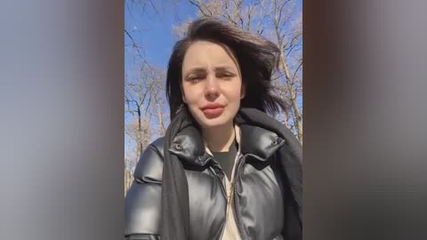 Media: Video of a young woman with fair skin, wearing a black puffy jacket, standing outdoors with bare trees and a clear blue sky in the background.