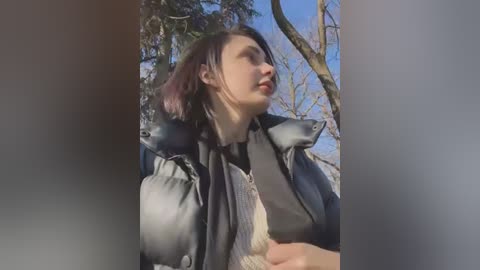 Media: Video of a young woman with fair skin, dark hair, and light makeup, wearing a black jacket and a beige sweater, holding a gray stuffed fox. She stands in a natural outdoor setting with bare trees and a blue sky in the background.