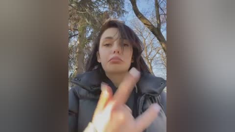 Media: A video of a young woman with long, dark hair, wearing a black puffy jacket, standing in a forest. She gestures with her hand, framed by bare trees and a clear sky.