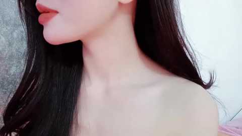 Media: A close-up video of a young woman with fair skin, long dark hair, and full lips, seen in profile. Her neck is bare, and the background is blurred, possibly a neutral-colored room.