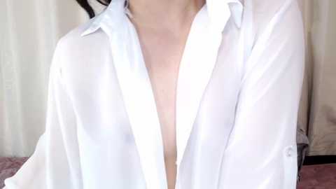 Media: Video of a light-skinned person wearing an unbuttoned white shirt, revealing their bare chest. Background features pale, vertical-striped curtains and a soft pink carpet. The image focuses on the upper torso, emphasizing the contrast between the white shirt and the skin.