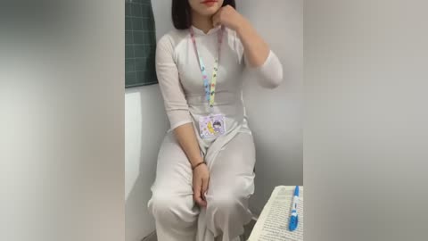 Media: Video of a woman with light skin and medium-length dark hair, wearing a white, ribbed, long-sleeved dress and lanyard with ID badge, sitting on a toilet, holding her chin thoughtfully.