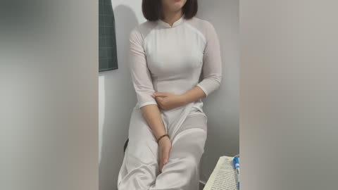 Media: Video of an East Asian woman with short black hair, wearing a white, form-fitting, long-sleeved dress. She sits with hands crossed on her lap in a small, dimly lit room with gray walls and a window with a green curtain.