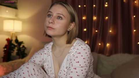 Media: A video of a young Caucasian woman with fair skin and a shoulder-length blonde bob, wearing a white blouse with small red floral patterns, seated in a cozy room with warm lighting and string lights.