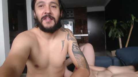 Media: Video of a shirtless man with a beard, tattoos, and long hair, sitting on a couch in a modern apartment with dark wooden furniture, a potted plant, and a blue sofa.