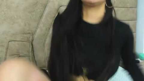 Media: Video of a woman with long black hair, wearing a black top, sitting on a beige car seat with a blurred background.