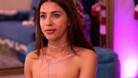 Media: Video of a young, attractive woman with long, dark hair, wearing a silver choker necklace, seated in a modern living room with blue furniture and vibrant lighting.