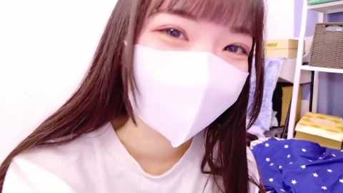 Media: Video of a young Asian woman with long, straight brown hair, wearing a white mask, white t-shirt, and blue-patterned pajama bottoms, in a cluttered room with shelves and clothes.