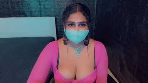 Media: Video of a South Asian woman with medium skin tone, wearing a pink top revealing cleavage, turquoise face mask, and braid, seated in a dimly-lit room with brick walls.