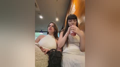 Media: Video of two women in a modern room, one in a white halter top, the other in a white dress, sipping from a pink bottle.