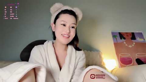 Media: Video of a smiling East Asian woman with fair skin, wearing a fluffy white robe, in a cozy room with a green wall and a poster featuring a woman in a swimsuit.