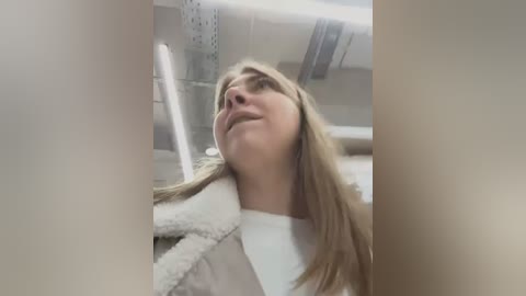 Media: Video of a blonde woman with a serene expression, wearing a white robe, inside a bright, sterile medical room with metallic and white walls.