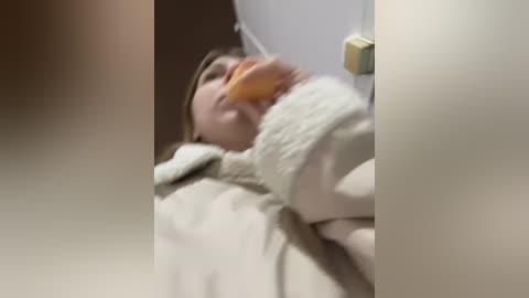 Media: A video of a young woman lying on a bed, wearing a beige robe, holding a large orange object, with a blurred background.