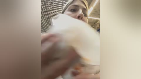 Media: A video captures a close-up of a woman's face and hand holding a piece of food, with a blurred background of a tiled ceiling and beige walls.