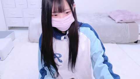 Media: A video of an Asian woman with long black hair, wearing a pink mask, white tracksuit with blue sleeves, and sitting on a white bed in a minimalist, white room.