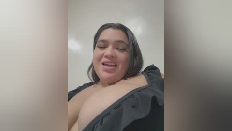 Media: Video of a plus-sized Latina woman with medium skin tone, short black hair, wearing a black top that reveals her large breasts, smiling, with a blurred background.