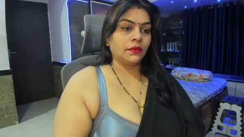 Media: Video of a South Asian woman with medium skin tone, long dark hair, wearing a light blue spaghetti strap top and black shawl, sitting on a gaming chair in a dimly lit bedroom with a bed, curtains, and bookshelf in the background.