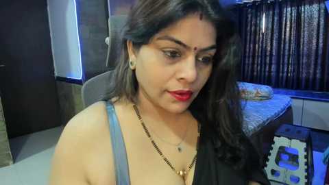 Media: Video of a South Asian woman with medium skin tone, long dark hair, red lipstick, wearing a black top and gold necklace, sitting indoors with a bed and window in the background.