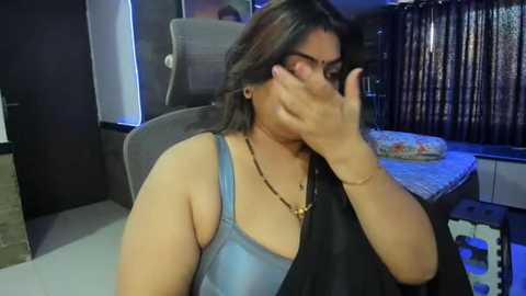 Media: Video of a South Asian woman with medium skin tone, wearing a blue tank top and black shawl, sitting in a dimly lit room with a grey chair and a bed with patterned bedding.