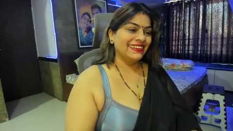 Media: Video of a smiling South Asian woman in a blue tank top, with dark hair, in a modern bedroom with a bed, curtains, and artwork.