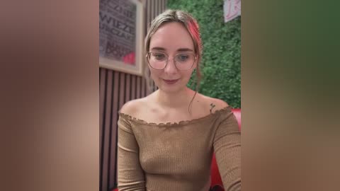 Media: Video of a young Caucasian woman with fair skin, glasses, and a brown off-the-shoulder top, standing in front of a wall with a green leaf pattern and black and white text, smiling softly.