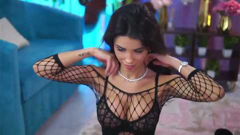 Video of a brunette woman with medium breasts, wearing a black fishnet top and pearl necklace, adjusting her hair, in a stylish, brightly lit room.