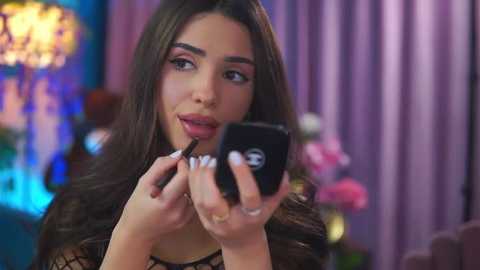 Video of a young woman with long, dark hair, applying makeup with a black lipstick applicator, in a brightly lit room with blurred, colorful background.