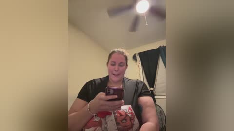 Media: Video of a smiling woman with light skin, brown hair tied back, wearing a black shirt and gray cardigan, holding a smartphone and a plush toy in a beige room with a ceiling fan and window with black curtains.