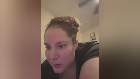 Media: Video of a young woman with light skin and brown hair pulled back, wearing a black shirt, looking introspective. Background shows beige walls, a ceiling fan, and a framed picture.