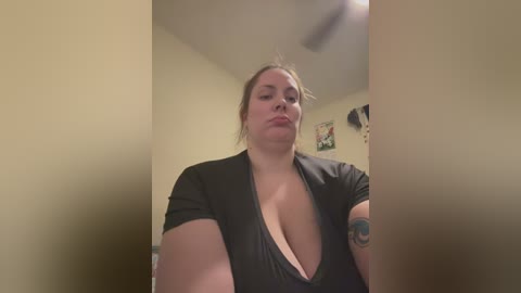 Media: Video of a plus-sized woman with light skin, wearing a black top that reveals cleavage, standing in a beige room with posters on the wall.