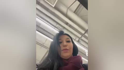 Media: Video of a young woman with long, dark hair, wearing a burgundy hoodie, taken from a low angle inside a fluorescent-lit, industrial building.