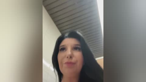 Media: Video of a young woman with long black hair and light skin, wearing black makeup, standing in a modern, minimalist hallway with a ribbed ceiling and light walls.