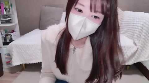 Media: Video of an East Asian woman with long brown hair, wearing a white face mask and off-white sweater, kneeling on a beige carpet in a room with a white bed and shelves filled with toiletries.