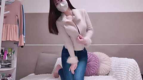 Media: Video of a slim, light-skinned woman with long brown hair, wearing a white cardigan, blue jeans, and a white face mask, kneeling on a bed with pink and white pillows, in a tidy, beige-walled bedroom.