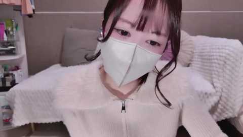 Media: Video of an Asian woman with straight black hair, wearing a white ribbed sweater and a white surgical mask, lying on a white bed in a dimly lit room.