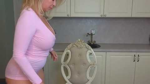 Media: Video of a blonde, fair-skinned woman in a tight pink long-sleeve top, showing a large bust, standing in a modern kitchen with white cabinets and a beige tufted chair.