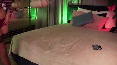 A video of a modern, dimly-lit bedroom with a large bed, beige sheets, a remote control, and green ambient lighting.