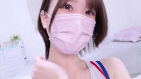 Media: Video of an Asian woman with shoulder-length brown hair, wearing a pink surgical mask, and a white sleeveless top with blue and red stripes, standing in a bright, minimalist room with white walls and a bed.