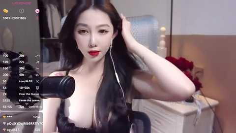 Media: Video of an East Asian woman with long black hair, fair skin, and red lipstick, wearing a black top, holding a microphone in a dimly lit room with a bed, white dresser, and window.