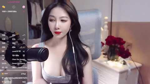 Media: Video of an East Asian woman with long black hair, wearing red lipstick, sitting in a dimly lit room with a microphone, flowers, and a white dresser in the background.