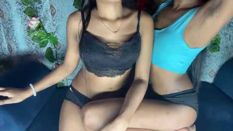 Media: Video of two young women with medium skin tone, one in black lace bralette and shorts, the other in blue tank top and shorts, sitting on a couch with floral-patterned curtains in the background.