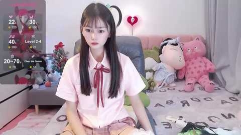 Media: Video of an Asian woman with long black hair and fair skin, wearing a pink school uniform, sitting on a bed with plush toys and a TV screen displaying a game in the background.